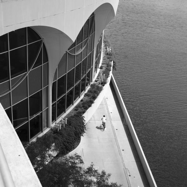 View from the top of Monona Terrace looking down towards the large arched windows. There are plants and trees in the concrete planters that are part of the architecture of the building. On the bike and pedestrian path along Lake Monona is a person riding a bicycle.