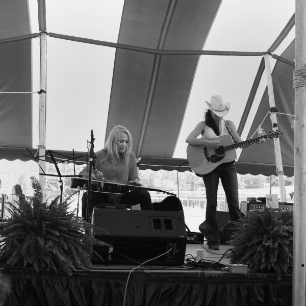 Shannon McNally is standing on a stage under a tent at the annual La Fete de Marquette Festival, playing guitar and singing roots and blues songs. She is accompanied by Cindy Cashdollar on the Dobro guitar. 