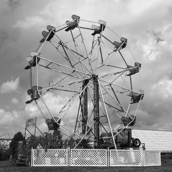 View across lawn towards a woman standing behind a fence at the base of the Ferris wheel waiting for customers at the La Fete de Marquette Festival. 