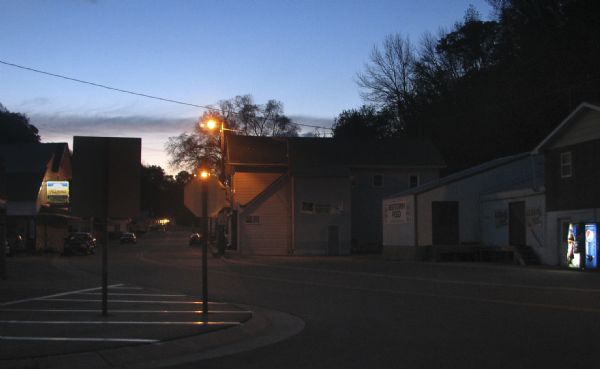 Dusk in downtown Beetown. View down curved street towards an illuminated Hamms sign on a building along the block on the left. A sign that reads "Beetown Feed" is on a building on the right.