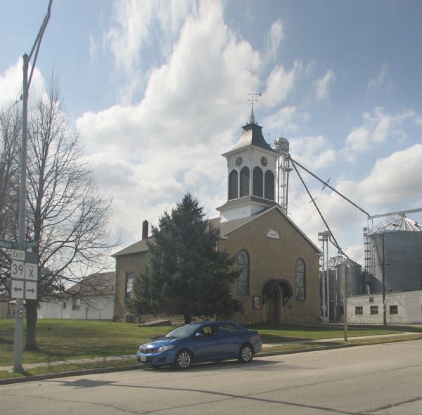 A stone church built in 1851 located at the junction of Highway 39 and County Trunk X. There is a large tree next to the church and a car parked on the street in front.