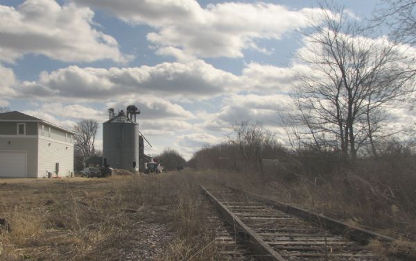 A view down train tracks overgrown with weeds. A building and metal silo are in the background.