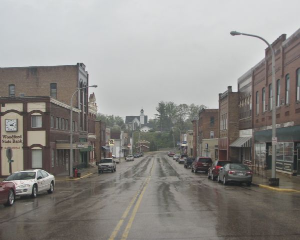 A view of the downtown area on a rainy day. The Woodford State Bank is on the left, and the street is lined with various storefronts and taverns. There is a church in the distance.