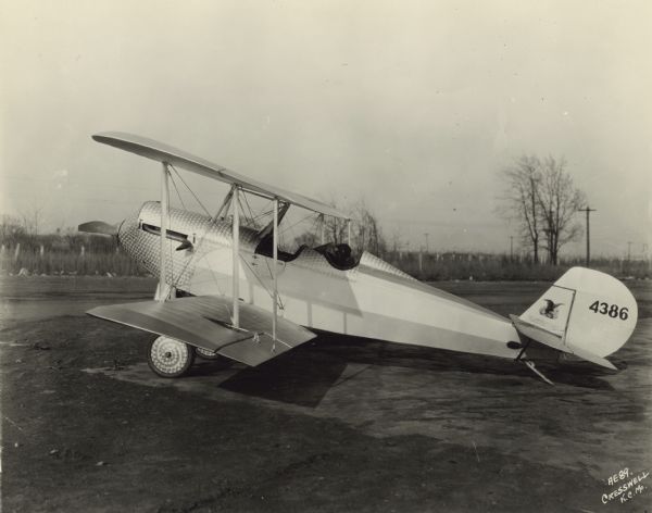 Left side view of an American Eagle Model A-101, a light 2/3-seat biplane, sitting in a field. There is a man sitting in the rear cockpit wearing an aviator cap and goggles. The tail identifier reads: "4386." The airplane body has a distinctive shiny steel texture with a circular pattern around the nose and the cockpits, and on the two front wheels.