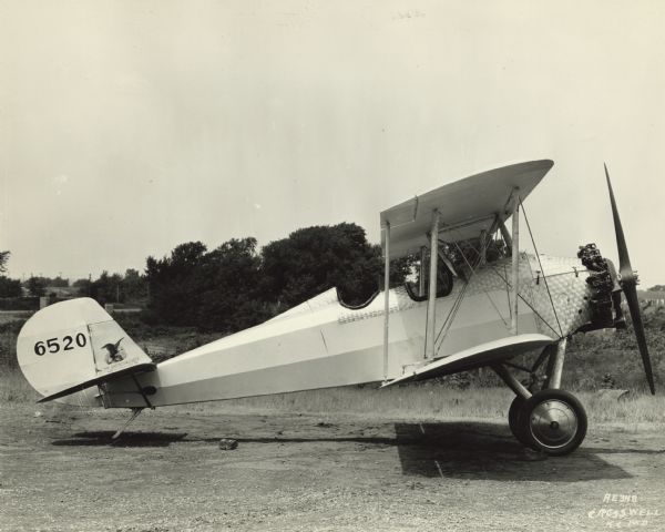 Right side view of an American Eagle Model A-129 sitting in a field. The tail identifier reads: "6520." The airplane body has a distinctive shiny steel texture with a circular pattern around the nose and the cockpits.