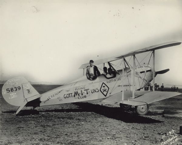 Right side view of an American Eagle Model A-101, a light 2/3-seat biplane, sitting in a field. The tail identifier reads: "5839." Four men are posing with the airplane: the pilot is sitting in the front cockpit, wearing an aviator cap and goggles, with another man sitting beside him on the left. The third man is sitting in the rear cockpit, and the fourth man is standing along the side of the airplane behind the three men. A number of slogans have been painted onto the side of the airplane: "Air Male," "Excuse Our Backwash," "In God We Trust," "Flying GOTT M*I*T UNS Club," "All Hope Abandoned Ye Who Enter Here," "Business End," "Save Old Iron Sides," "Engine Room." There is also a drawing of a skull and crossbones. The airplane body has a distinctive shiny steel texture with a circular pattern around the nose and the cockpits.