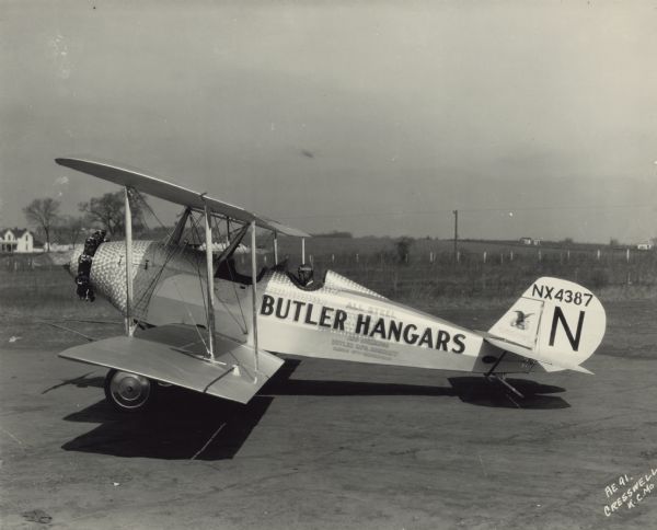Left side view of an American Eagle Model A-129, a three-seat open-cockpit sports/utility biplane, sitting on a runway. A man is sitting in the rear cockpit wearing an aviator cap and goggles. The tail identifier reads: "NX4387." An advertisement for Butler Manufacturing Company is printed on the side of plane and reads: "All Steel Butler Hangars And Buildings Butler Mfg. Company Kansas City — Minneapolis." The airplane is powered with a quick radial air-cooled 120 hp engine. The airplane body has a distinctive shiny steel texture with a circular pattern around the nose and the cockpits.
