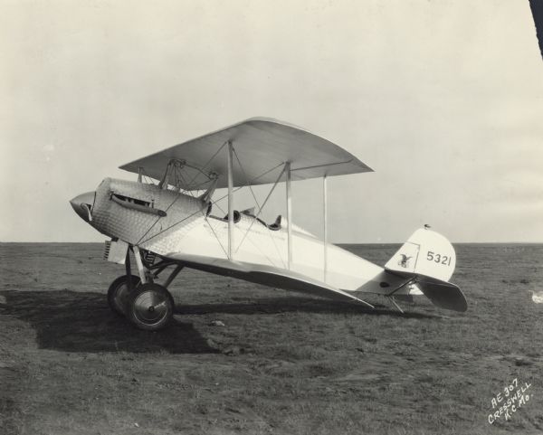 Left side view of an American Eagle Model A-101, a light 2/3-seat biplane, sitting in a field. The tail identifier reads: "5321." The airplane body has a distinctive shiny steel texture with a circular pattern around the nose and the cockpits.