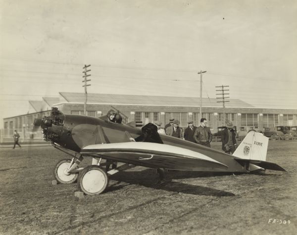 Side view of a Fairchild Model 21 sitting on a runway with two people wearing aviator hats and goggles sitting in the front cockpit, and one person standing near the wing. In the background are six men are standing and examining the airplane. The tail identifier reads: "X139E."