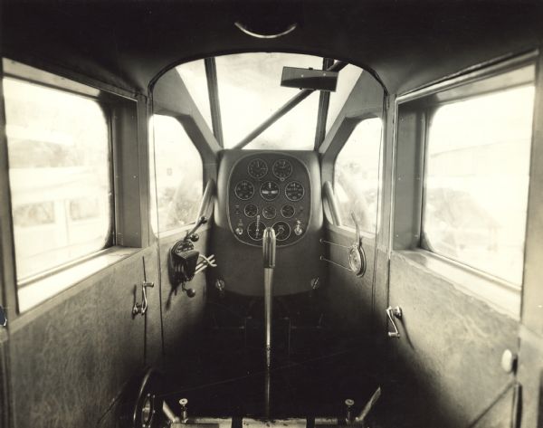 Interior forward view of a Fairchild 71 cabin showing instrument board, engine controls, and control stick. The mirror over the instrument board allows the pilot to read the compass located above his head without looking up. The figures on the compass are reversed to allow for reading in the mirror. The lever on the far right regulates air circulation to cool the engine. In the bottom left corner is a wheel for stabilizer adjustment.