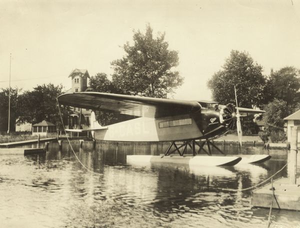 Three-quarter view of front right side of a Fokker Super Universal seaplane floating in the water near a dock. Powered with Pratt & Whitney 410 hp Wasp motor. Designed with full cantilever wing and two metal floats.