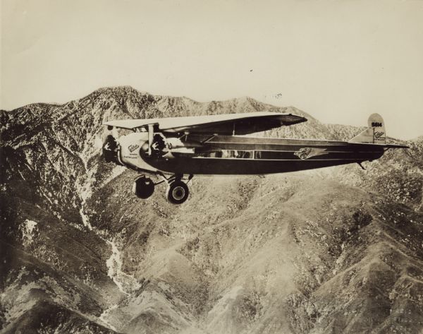 Left side aerial view of a Fokker Trimotor F-10 'Deluxe' in flight with mountains in the background. The airplane was owned by Richfield Oil. The tail identifier reads: "5614."