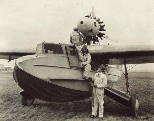 Three-quarter view from front left of a Fokker Amphibian, Model F-11, sitting on a runway. Two men are sitting on the aircraft, and one man is standing on the ground next to the door. The men are wearing Fokker coveralls. This seaplane is equipped with a 425 hp Pratt & Whitney Wasp engine and retractable landing gear sponsons.