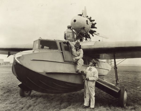 Three-quarter view from front left of a Fokker Amphibian, Model F-11, sitting on a runway. Two men are sitting on the aircraft, and one man is standing on the ground next to the door. The men are wearing Fokker coveralls. This seaplane is equipped with a 425 hp Pratt & Whitney Wasp engine and retractable landing gear sponsons. 