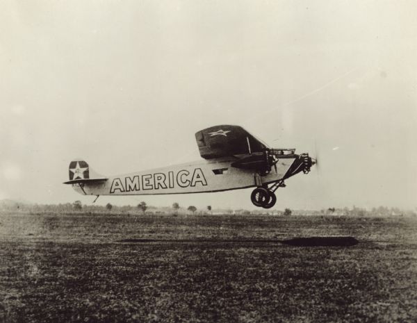 Right side view of a Fokker C-2 Trimotor monoplane named "America" in flight just above a field. The airplane was equipped with three 220hp Wright J-5 engines. The tail identifier reads: "NX 206." "America" was flown by pilot Richard Byrd from New York to France in June of 1927 shortly after Charles Lindbergh won the Orteig Prize for completing the same trans-Atlantic flight. "America" was unable to land in Paris due to heavy fog and instead was forced to land just off the coast of Normandy, resulting in the destruction of the plane.