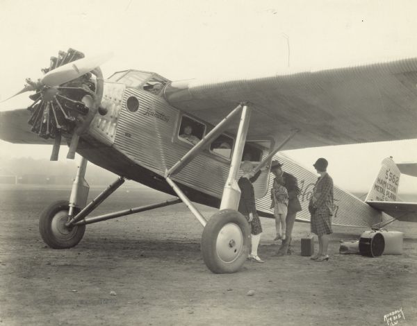 Three-quarter view of a Hamilton Metalplane H-21 sitting on a runway. The airplane was equipped with a 400 hp Pratt & Whitney engine. The tail identifier reads: "5562." The pilot is sitting in the cockpit, and three women are looking out of the side windows. On the ground a man is holding up a young child near the open side door, and two women stand nearby. Pieces of luggage are on the ground. "Hamilton" and "Hamilton Metalplane Co. Milw(aukee)" are painted on the side of the airplane and the tail.