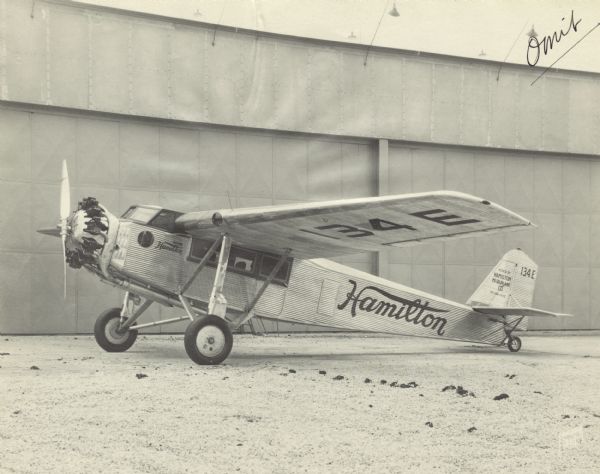 Right side view of a Hamilton Metalplane H-47 parked near a large building. The airplane is equipped with a 525 hp Pratt & Whitney Hornet engine. The tail and wing identifier reads: "134 E." "Hamilton" is painted on the side, and "Mfr'd by Hamilton Metalplane Co. Milwaukee" is on the plane's tail.