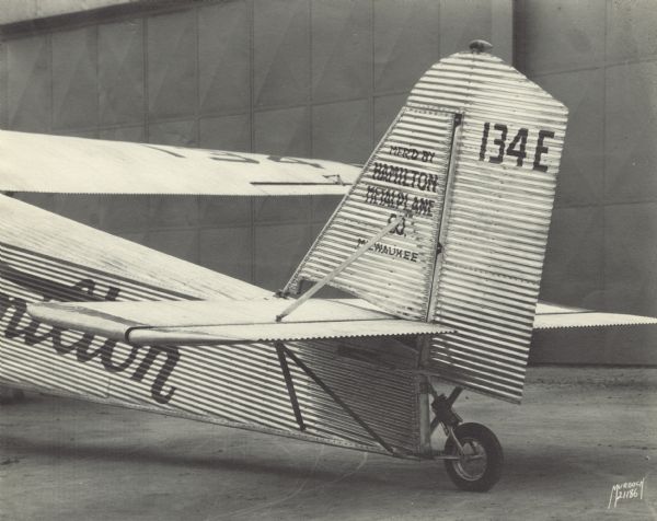 Three-quarter from left rear of the tail end of a Hamilton Metalplane H-47 parked near a large building. The tail identifier reads: "134 E." The airplane is equipped with a 525 hp Pratt & Whitney Hornet engine. Painted on the tail is: "Mfr'd by Hamilton Metalplane Co. Milwaukee."