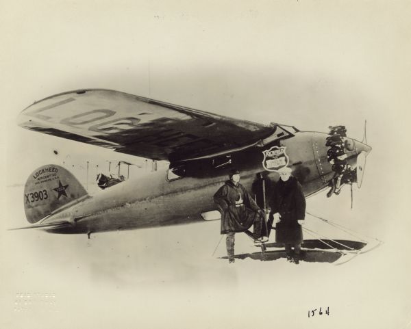 Right side view of a Lockheed Vega 1 sitting in the snow. The tail identifier reads: "X3903." The airplane is equipped with a 220hp Wright J-5 engine. Captain George Hubert Wilkins and Ben Eielson are standing beside the airplane. This photograph was taken just prior to the completion of the first Arctic crossing by air in April 1928. The journey took approximately 20 hours and covered a distance of 2,200 miles.