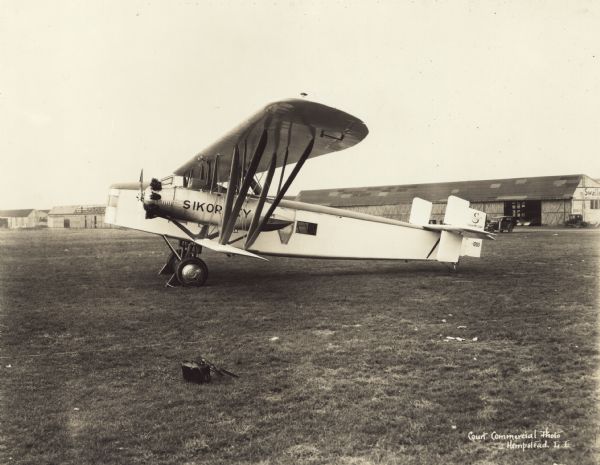 Left side view of a Sikorsky S-37-1 Guardian sitting on a runway. The tail identifier reads: "1283."There are automobiles parked near buildings in the background, and a sign on a building on the right reads in part: "Swallo[w], American's First Comm ..." 