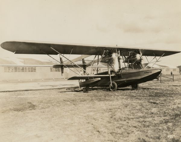 Three-quarter view from front right of a Sikorsky S-38, an amphibious plane equipped with two Pratt & Whitney Wasp engines, sitting on a runway. Painted on the side of the plane is the name: "Liberty." In the background is a long industrial building.