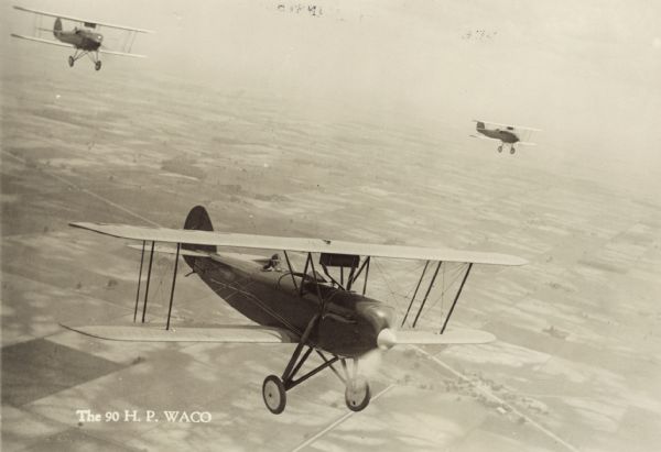Aerial view of a WACO 10 (sometimes referred to as WACO 90 because of its horsepower) in flight, with two other WACO planes trailing in the background. Fields, roads, and houses are below in the background. The airplane is powered by a 90hp Curtiss OX-5 engine.