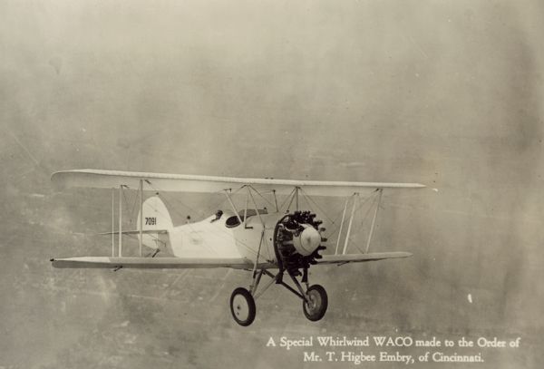 Aerial view of a WACO ASO (also known as a J-5 Straightwing and Whirlwind Waco) in flight. Below in the background is a town and fields. The tail identifier reads: "7091." The airplane is equipped with a 220 hp Wright J-5 engine. It was made to the order of T. Higbee Embry from Cincinnati. The airplane was ultimately destroyed in April, 1991 near Zellwood, Florida while being used to observe alligators. The plane dove too close to the water and was unable to pull up at such a low altitude. All three occupants were killed.