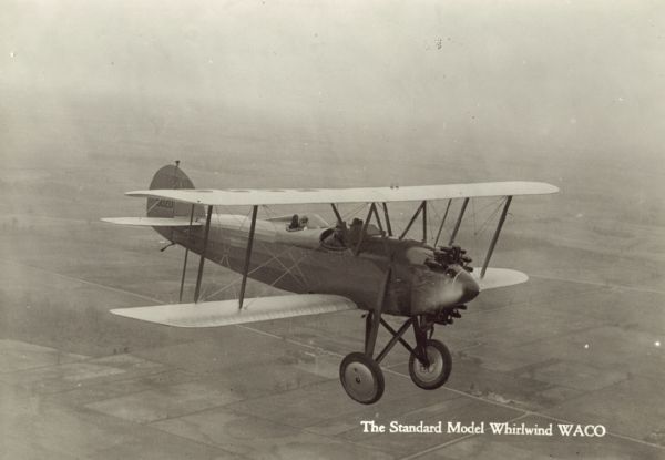 Aerial view of a WACO ASO (also known as J-5 Straightwing and Whirlwind Waco) in flight. Two men are sitting in the front open cockpit, and one man is sitting in the rear open cockpit. The airplane is equipped with a 220 hp Wright J-5 engine. Fields, roads, and houses are below in the background.