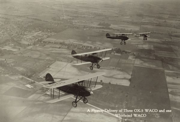 Aerial view of four WACO biplanes in flight. They are being delivered from the manufacturing company in Ohio. The three airplanes in the foreground are WACO 10 biplanes powered by 90hp Curtiss OX-5 engines. The airplane in the far background is a WACO ASO biplane powered by a 220 hp Wright J-5 engine. The wing identifier on the far left plane reads: "3287." Below in the background below are fields, roads, and houses.