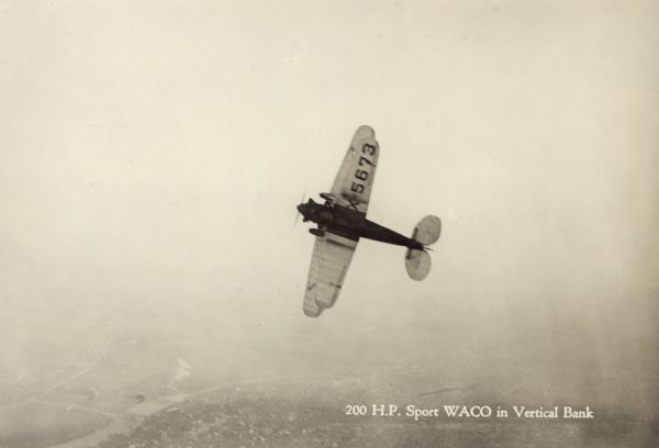 Aerial view of a WACO ATO (also known as 10-T Taperwing) in flight during vertical bank. The wing identifier reads: "5673." The airplane is powered by a 220hp Wright J-5 engine. Below in the background is a river, fields, and a town.
