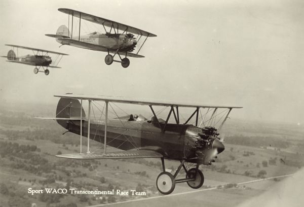 Aerial view of three WACO ATO (10-T Taperwing) biplanes in flight. The wing identifiers of the two planes furthest to the right read: "7527" and "5673" (left to right). The airplanes are powered by 220hp Wright J-5 motor engines. They were flown in 1928 as part of a transcontinental air race from New York to Los Angeles. Fields and roads are below in the background.