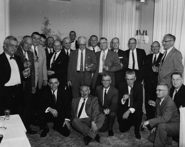Group portrait of Gisholt Machine Company office workers in formal attire. Most of the men are standing and holding drinks in their hands. A few are kneeling on the floor in front, and Sid Boyum (center, sitting on the floor) has a cigar in his mouth.
