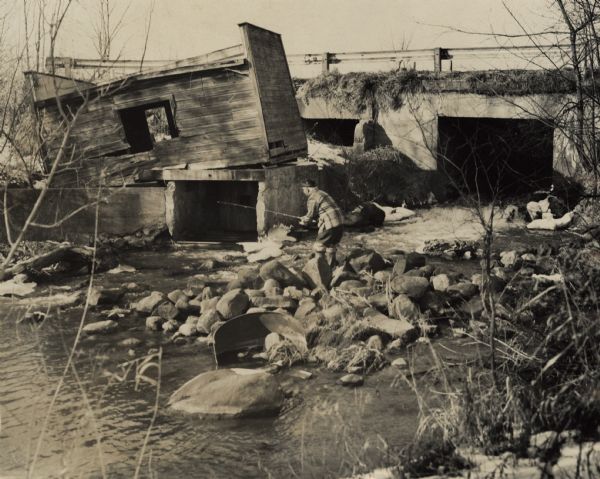 View from shoreline towards Sid Boyum fishing while standing on a pile of rocks near a river. Near Sid on the opposite shoreline is a collapsed building on a foundation, and in the background is a bridge over the river.