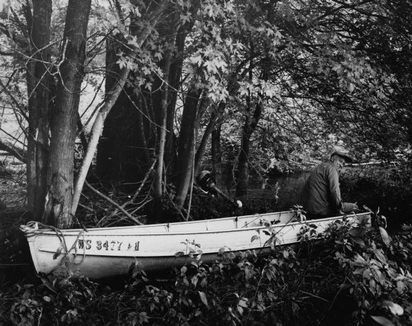 Man sitting at the aft of a canoe in a wooded river area. There is an outboard motor leaning against a tree in the center of the image. Handwritten text captioning the photograph reads: "Portaging - White River near Fox River."