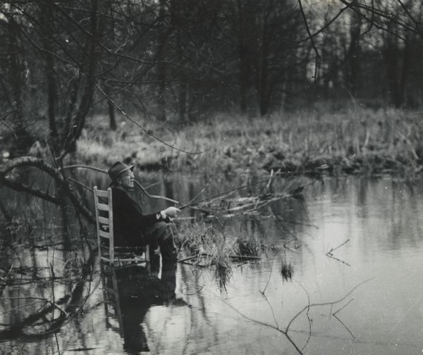 View across a marshy area towards a man, Mike Heberlein (of Cambridge), fishing while sitting in a wooden, ladder-back, dining chair that is submerged in the water. Wetlands are seen in the background.