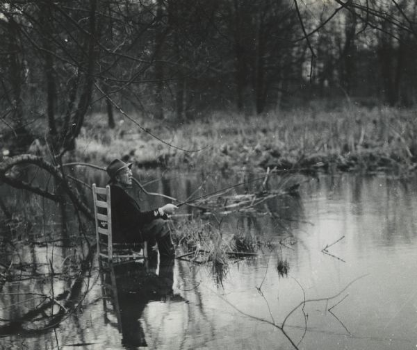 View across a marshy area towards a man, Mike Heberlein (of Cambridge), fishing while sitting in a wooden, ladder-back, dining chair that is submerged in the water. Wetlands are seen in the background. 