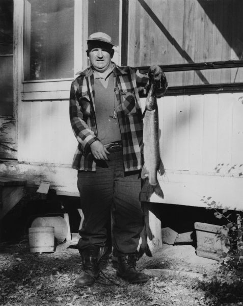 Full-length portrait of Sid holding a northern pike on a stringer in front of what may be a cabin.