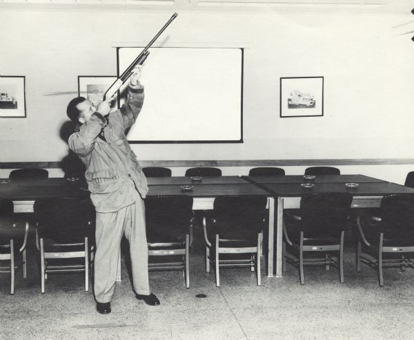 View of a man posing and aiming a gun into the air inside a Gisholt conference room. The man is wearing a hunting jacket and he is standing in front of chairs along a long table set with ashtrays.