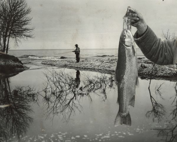 Composite photograph of one forming the background that shows a man with a pipe in his mouth, fishing at the edge of an inlet of a lake and stream, and of the other with a man's arms holding a fish on a stringer in the foreground. This image appeared in the <Wisconsin State Journal</i>'s section of "Outdoor World" on March 18, 1973 along side an article and portrait of Sid.