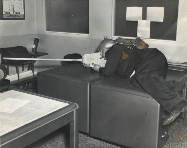 Sid, with a cigar in his mouth, a metal lampshade on his head, and wearing a backpack, is laying across two desks in an office with a broom in his arms as if he is holding a gun. Sid's nails are likely stained with pyrogallol, a staining agent used in the development of film. 