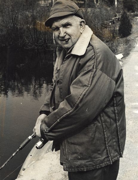 Three-quarter length portrait of Sid Boyum holding a fishing rod and wearing a coat and hat, with a cigar in his mouth. He is standing on a path above a river or stream. Behind him is a wooded shoreline, and a building on a hill.
