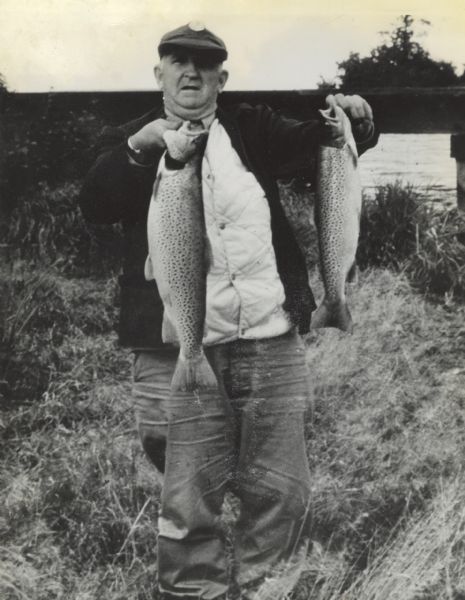 Portrait of Sid Boyum holding two female brown trout, one 8.5 pounds and the other 6 pounds. In the background is a bridge or pier, and a body of water.