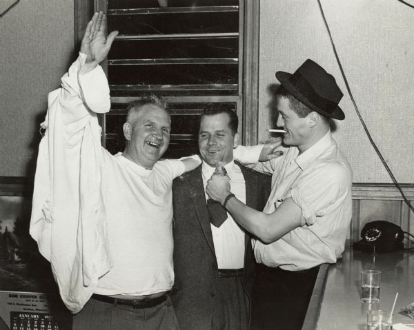 Group portrait of Sid Boyum standing with two men inside a bar. Sid, on the left, has his arm up in the air, and his button-down shirt appears to be torn, showing his undershirt. The man on the right is gripping the necktie of the man in the center. A calendar in the bottom left reads: "Bob Cooper Glass, January 1957."