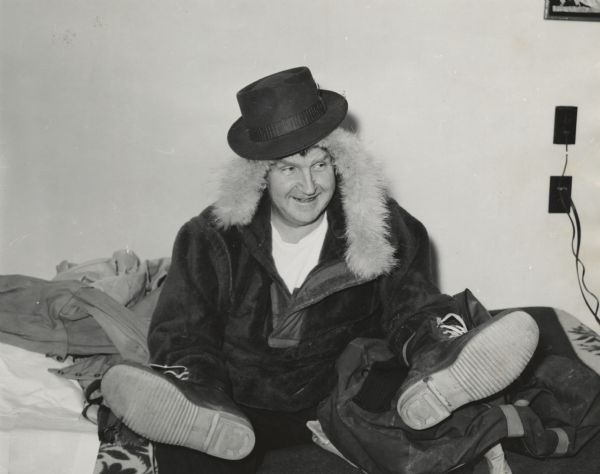 Portrait of Sid sitting on a bed with boots on his hands, and a coat and a hat. The fur-lining of his coat surrounds his face.
