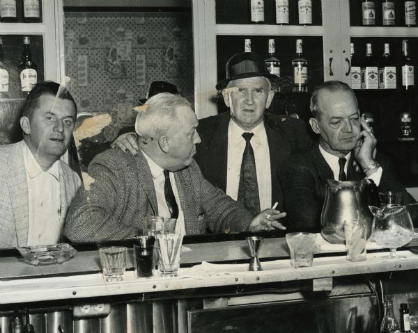 View from behind a bar of Sid, in center wearing a hat, standing with friends who are sitting at the bar. Sid's nails are stained, probably with pyrogallol, a staining agent used in the development of film.