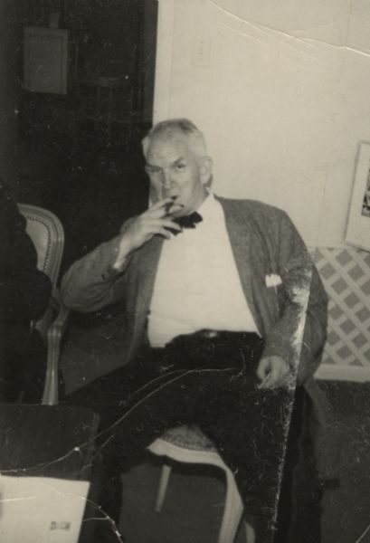 Sid Boyum sitting indoors in what appears to be a living room. He is wearing a bow tie and suit jacket. He is holding a cigar to his mouth.