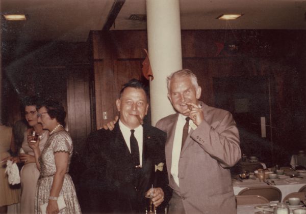 Portrait of Sid standing with another man at a gathering. Both men are wearing suits and have cigars in their mouths. Two women are in the background on the left. Behind them on the right are long dining tables.