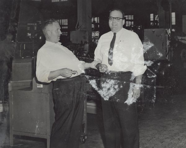 Sid and a colleague standing in what appears to be a work area. Sid is leaning back, while holding a pair of eyeglasses in his right hand and grasping his male colleague's right forearm. The man is holding a cigarette in his right hand. Both men are wearing white button-down shirts. A man is standing in the background on the left.