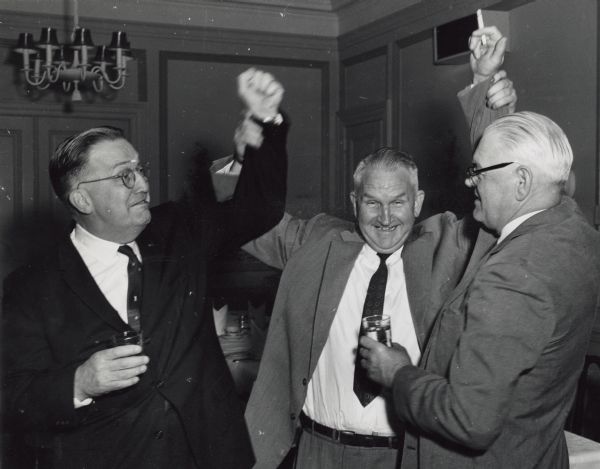 Sid standing between two Gisholt colleagues with his arms raised holding their right arms aloft. The two colleagues are holding drinks, and all three men are wearing suits. A chandelier and part of a place settings on a table are in the background.   