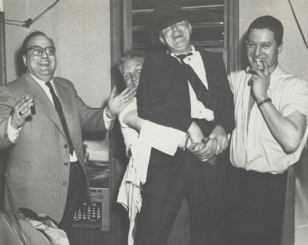 Sid posing for group portrait with three male colleagues. Sid is wearing a suit, tie, and a hat, and is being hoisted up by a colleague standing behind him. The man on the left is holding a drink, and the man on the right is smoking a cigarette. "Bob Cooper Glass Co., January 1957."